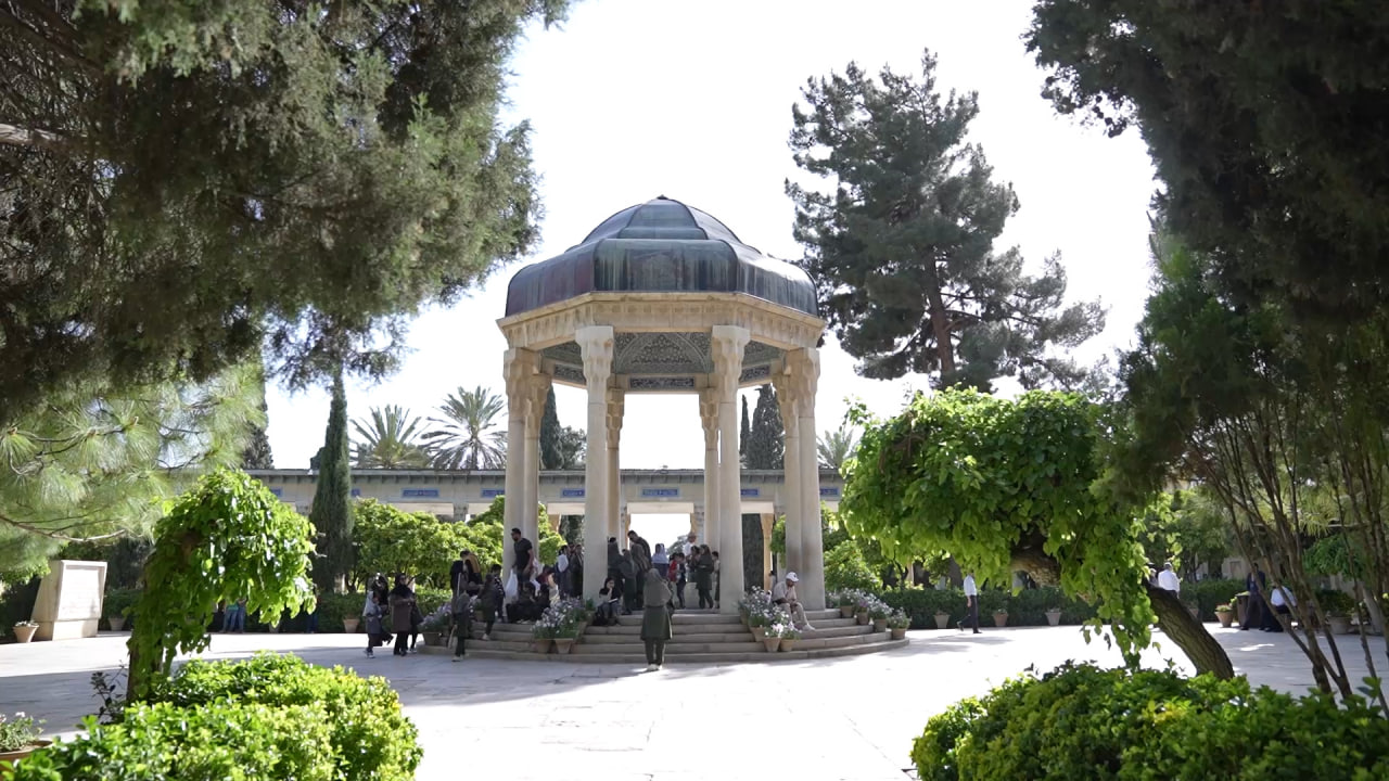 آرامگاه حافظ که از میان درختان اطراف نمایان میشود و افرادی در پسزمینه دیده میشوند. دوربین از پیشزمینه درختان حرکت میکند و به تدریج روی آرامگاه تمرکز میکند و ضمن نمایش مقیاس و فعالیت انسانی، جلوه بصری چشمگیر و فراگیری ایجاد میکند. این یکی از بهترین ویدیوها برای مستندهای فرهنگی، فیلمهای مسافرتی، پروژههای تاریخی، محتوای میراث ایرانی، داستانسرایی سینمایی و فیلمهای آموزشی است.