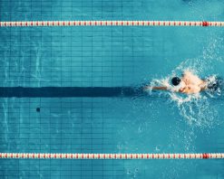 نمای هوایی از بالا، شناگر مرد در استخر شنا می‌کند. تمرین ورزشکار حرفه‌ای برای مسابقات قهرمانی، با استفاده از تکنیک کرال سینه و شنای آزاد. فیلم سینمایی با نمای عریض و آهسته و قابلیت ردیابی حرکت 4K